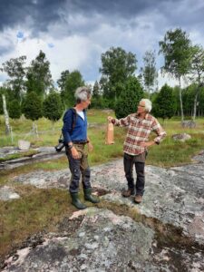 En man i rutig skjorta och grönsvarta byxor räcker över en papperspåse till en man i blå tröja och grönsvarta byxor. Det står på en berghäll mitt i en blomstrande äng.